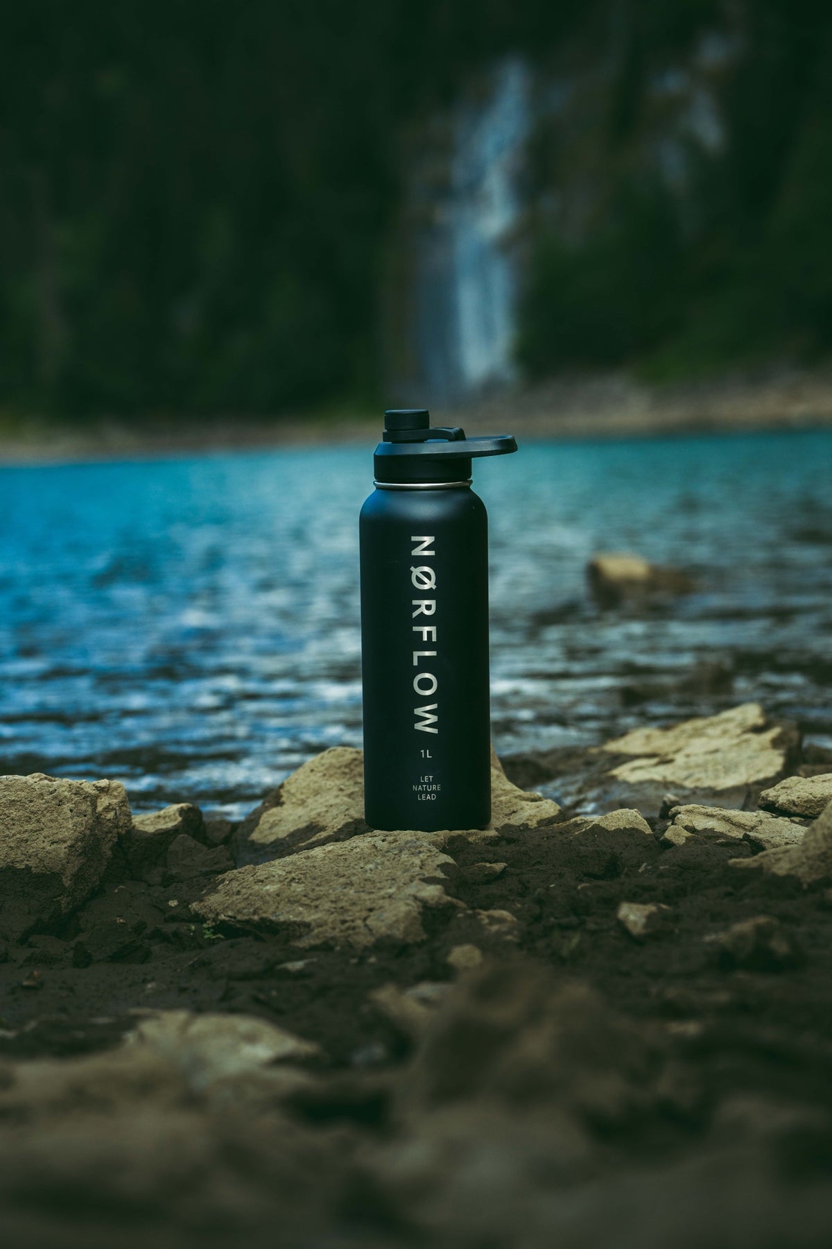 Botella térmica NORFLOW 1L negra sobre rocas junto a un lago con cascada