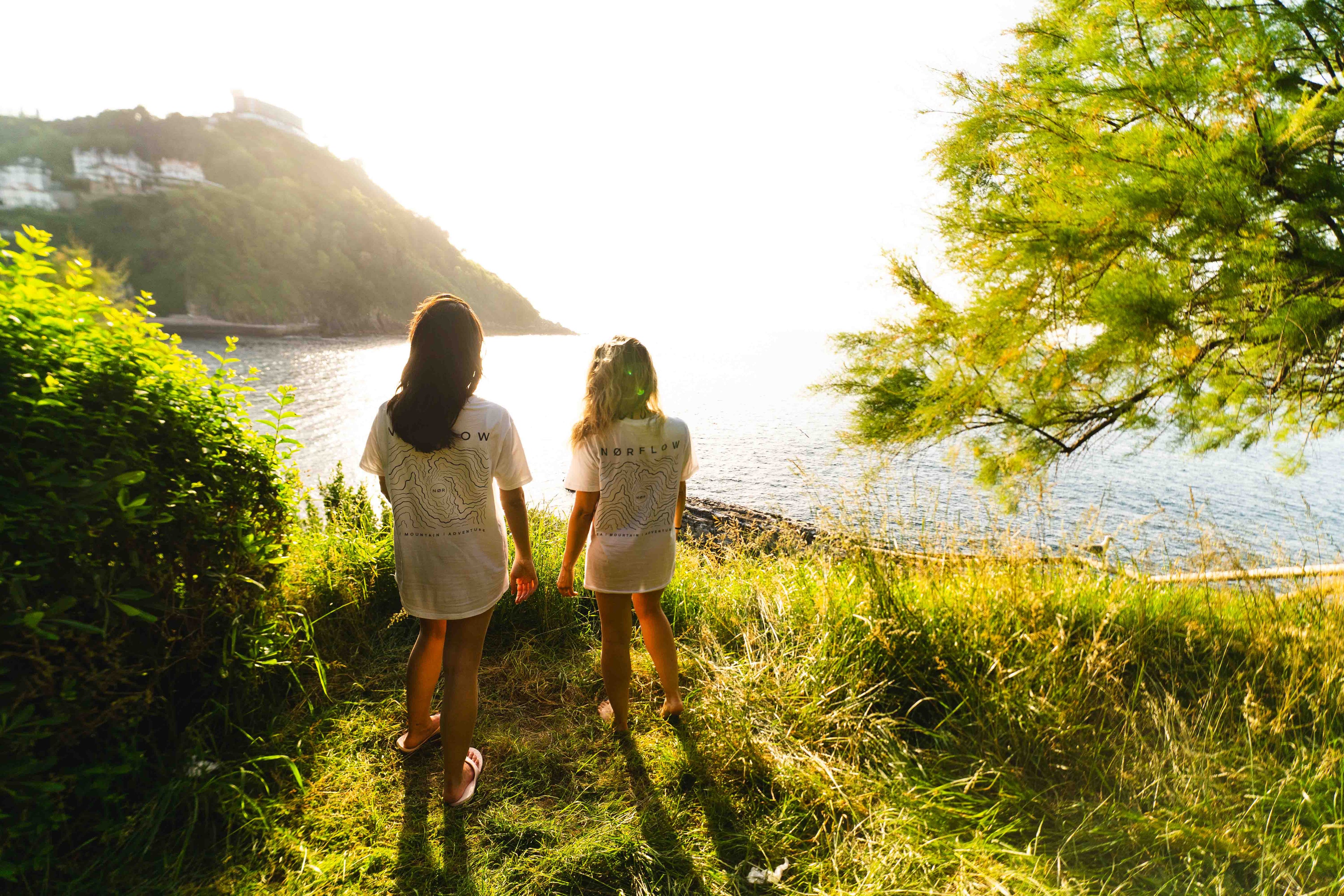 Dos jovenes en una isla usando la camiseta blanca de NØRFLOW  con un diseño de líneas de nivel de montaña. Ideal para actividades al aire libre y deportes acuáticos.Marca inspirada en el mar, la montaña y la aventura. Norflow nace de la necesidad de explorar y descubrir nuevos lugares perdidos por el norte. Diseña productos funcionales para acompañar experiencias al aire libre, combinando utilidad, durabilidad y un estilo de vida activo.
