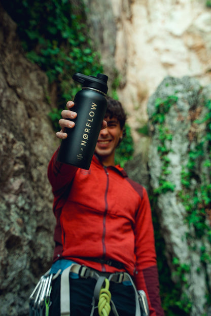 Escalador sonriendo con arnés y sudadera roja mientras muestra la botella térmica NORFLOW 1L