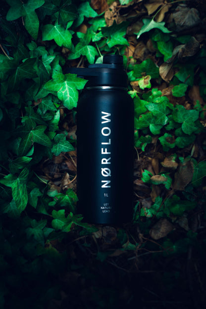 Botella térmica NORFLOW 1L negra entre hojas de hiedra en el suelo del bosque